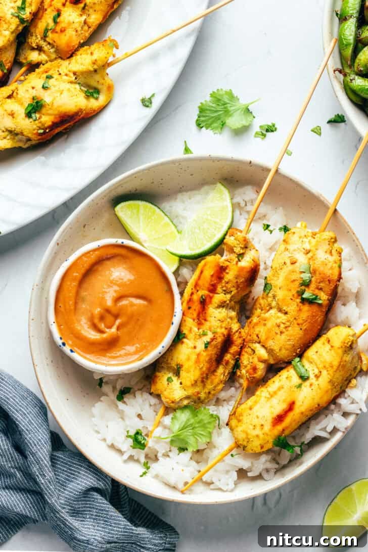 Sizzling Chicken Satay Skewers 4 Six pieces of chicken satay arranged on a white round plate with a side of fluffy white rice, vibrant lime slices, and a small white ramekin of rich peanut sauce.