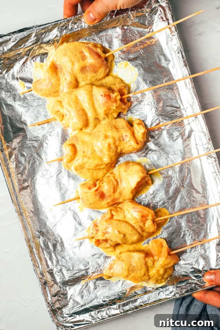 Sizzling Chicken Satay Skewers 8 A hand holding a baking sheet covered with aluminum foil, showcasing six raw, perfectly threaded chicken skewers, ready for grilling.