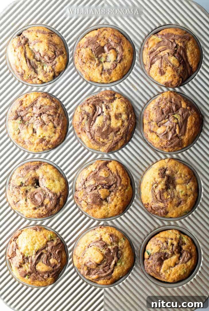 Decadent Zucchini Nutella Muffins 12 An inviting top-down view of a dozen perfectly golden-brown zucchini and Nutella muffins, still in their baking tin, showcasing their beautiful swirls and ready to be served.