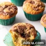 A close-up view of a dozen chocolate zucchini muffins with prominent Nutella swirls, presented in green cupcake wrappers. One muffin is thoughtfully split in half, revealing its moist interior and the beautiful marbling of the chocolate hazelnut spread.