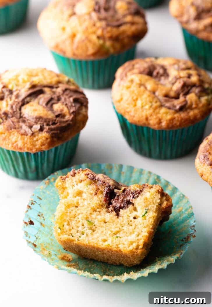 Decadent Zucchini Nutella Muffins 14 A vibrant display of a dozen freshly baked chocolate zucchini muffins, each adorned with a distinct Nutella swirl, perfectly arranged in green cupcake wrappers. One muffin is broken open to reveal its moist, tender interior and the enticing marbling of the chocolate and hazelnut.