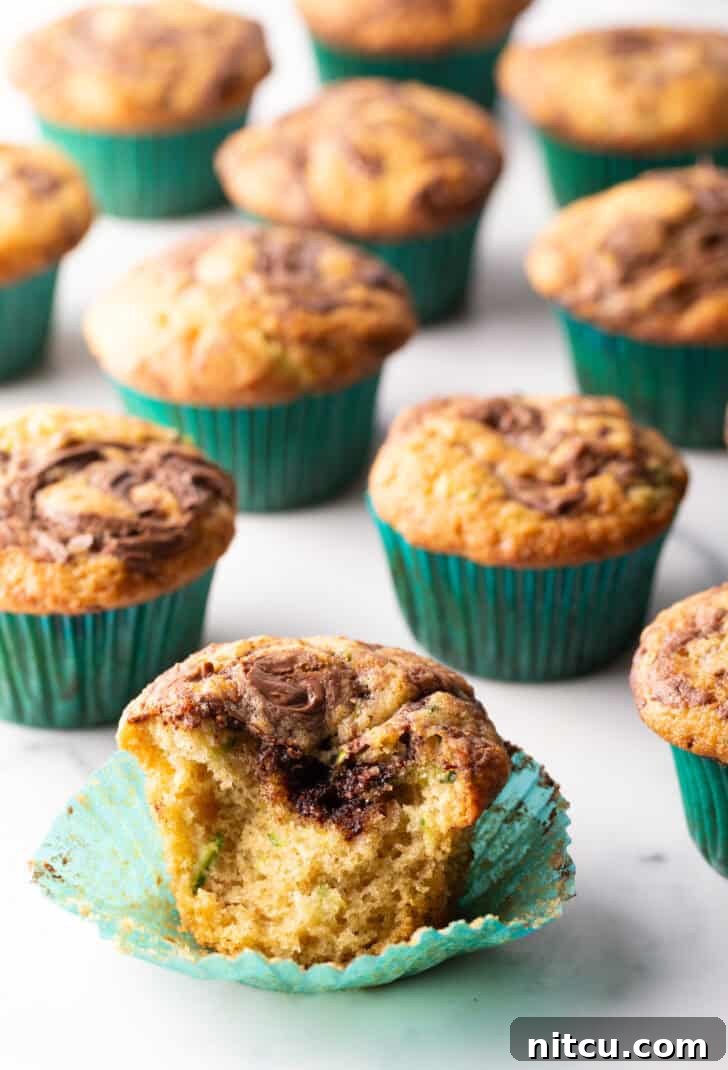 Decadent Zucchini Nutella Muffins 4 A close-up view of a dozen chocolate zucchini muffins with prominent Nutella swirls, presented in green cupcake wrappers. One muffin is thoughtfully split in half, revealing its moist interior and the beautiful marbling of the chocolate hazelnut spread.