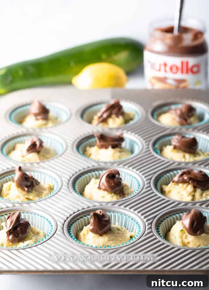 Decadent Zucchini Nutella Muffins 9 Unbaked zucchini muffin batter in a cupcake tin, each portion topped with a generous dollop of rich Nutella, poised for swirling.
