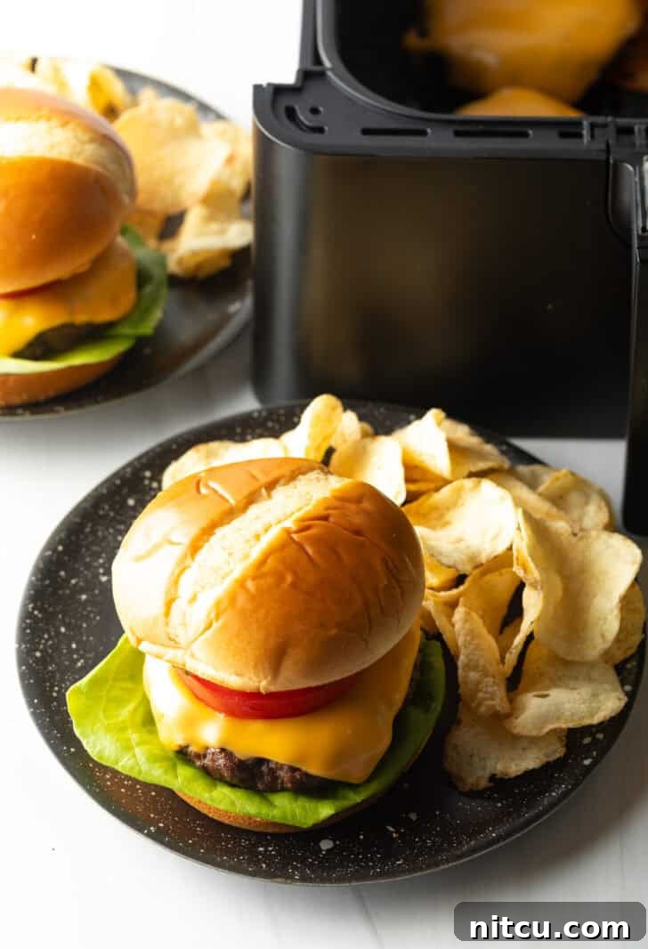 Aair fryer hamburger loaded with cheese, lettuce, tomatoes and pickle slices on a plate with potato chips.