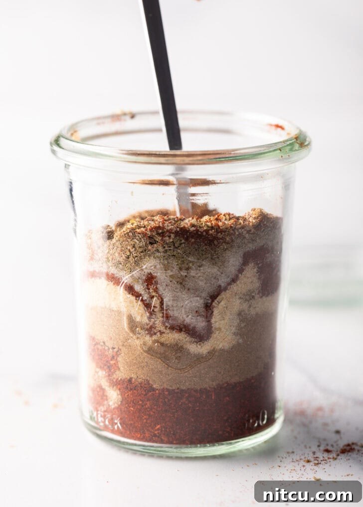 Zesty Chicken Taco Blend 7 A spoon actively stirring layers of chili powder, cumin, garlic powder, and other spices in a glass jar, demonstrating the mixing process.