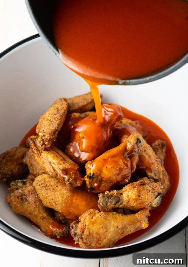 White mixing bowl with chicken wings, pouring buffalo sauce over the top.