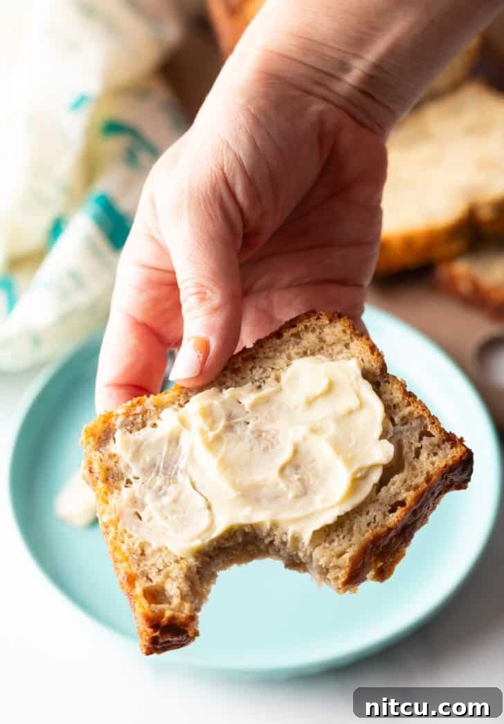 Effortless Banana Bread 15 A hand holding a moist slice of banana bread, generously slathered with butter, showing a bite taken from the corner.
