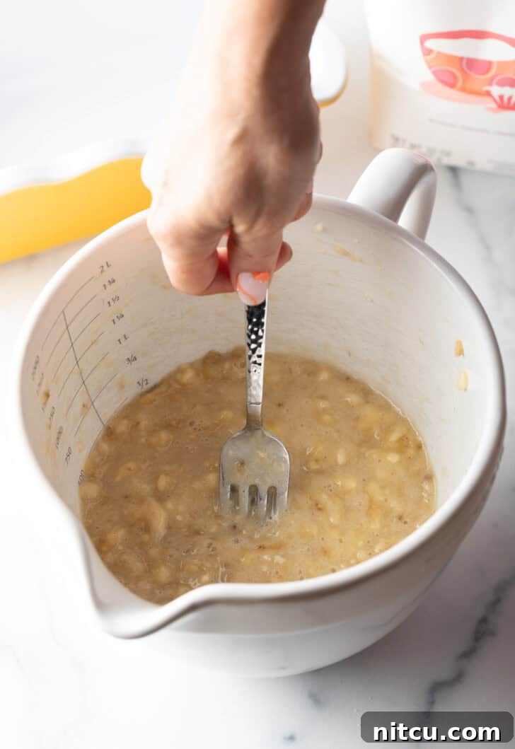 Effortless Banana Bread 7 A hand using a metal fork to mash ripe bananas in a white mixing bowl, illustrating the mashing process.