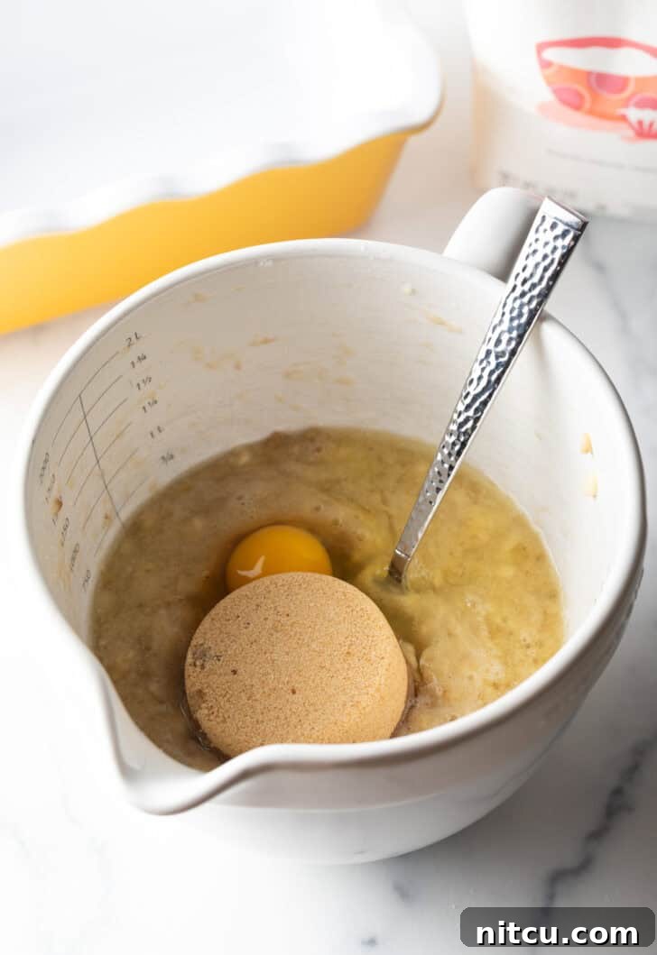 Effortless Banana Bread 8 Adding a scoop of brown sugar and a raw egg into a large white mixing bowl already containing mashed bananas, showing the next step in the recipe.