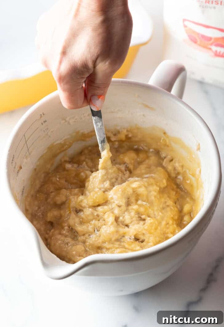 Effortless Banana Bread 9 A hand using a metal spoon to gently stir the banana bread batter in a large white mixing bowl, demonstrating the mixing technique.