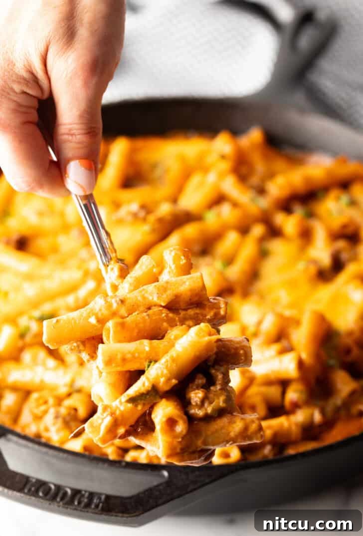 Homestyle Beef Bake 12 Hand using a large spoon to scoop noodle cheeseburger casserole from a large skillet towards camera. The casserole is steaming and freshly cooked.