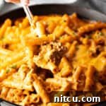Hand using a large spoon to scoop noodle cheeseburger casserole from a large skillet towards camera. A close-up of the finished dish.