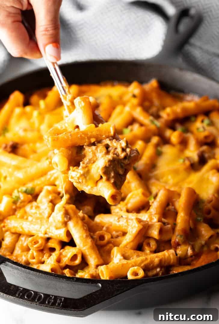 Homestyle Beef Bake 4 Hand using a large spoon to scoop noodle cheeseburger casserole from a large skillet towards camera. The rich, cheesy, and saucy texture is visible.