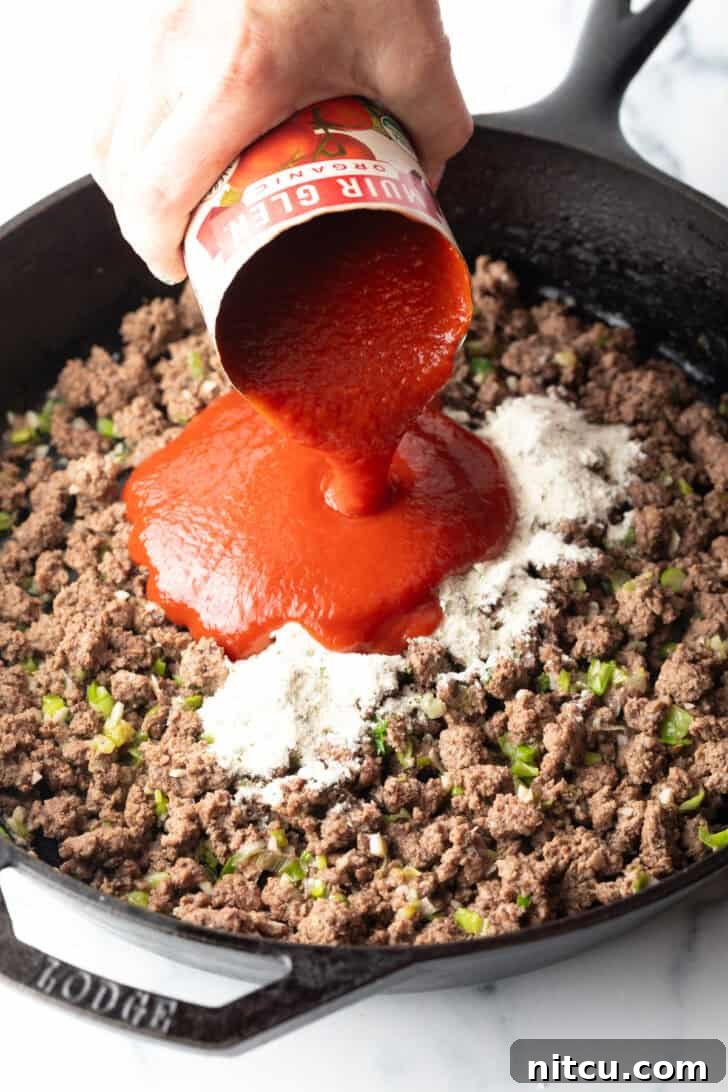 Homestyle Beef Bake 6 Hand adding canned tomato sauce to a skillet of cooked ground beef and ranch seasoning mix. The sauce is rich red, contrasting with the browned meat.