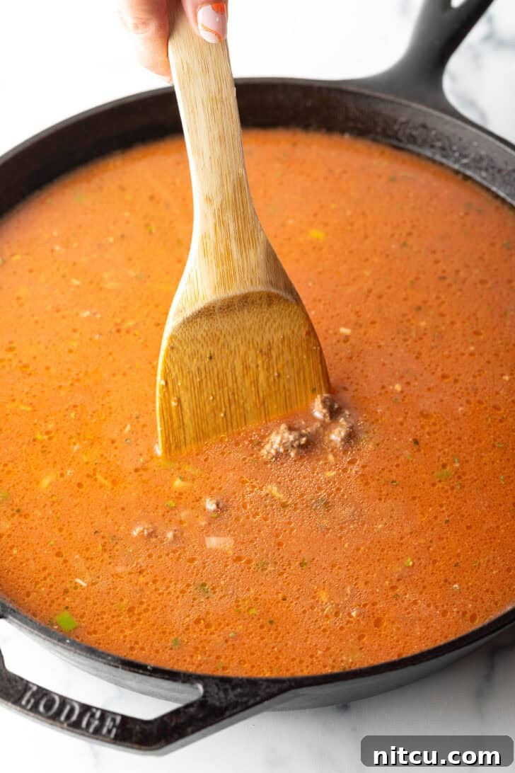 Homestyle Beef Bake 7 Using a wood spatula to stir tomato and beef mixture in a cast iron skillet. The mixture is simmering gently.