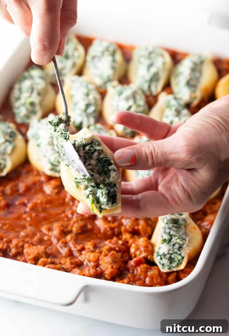 Hand using a metal spoon to fill a pasta shell with spinach and cheese filling.