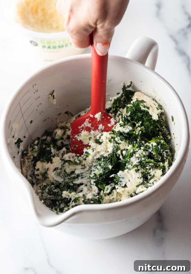 Using a red spatula to stir cheese and spinach in a large white mixing pitcher.