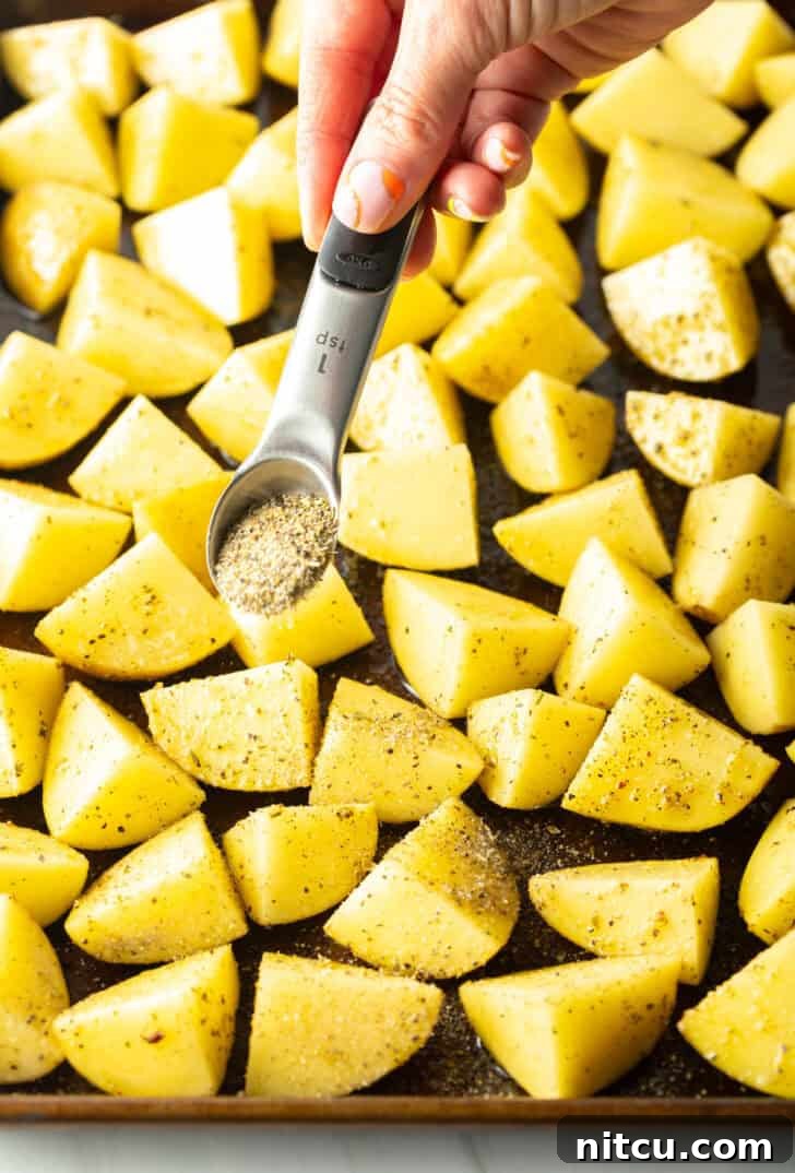 Crispy Potato Gold 7 Hand sprinkling seasonings from a small teaspoon overtop of potatoes chunks on a baking sheet.