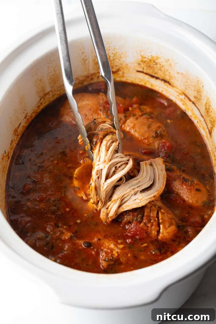 Fiesta Slow Cooker Chicken 10 Tongs holding up shredded chicken above a white slow cooker.