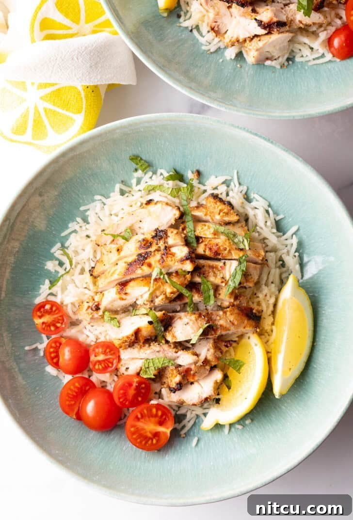 Shallow blue bowl with white rice topped with slices of marinated greek chicken, halved grape tomatoes, and two lemon wedges.