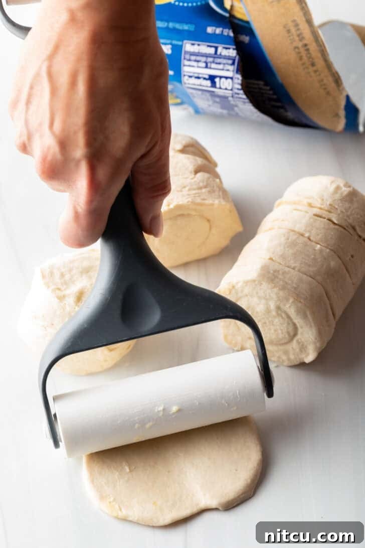 Effortless Slow Cooker Chicken and Dumplings 9 A hand using a rolling pin to press a raw biscuit flat on a cutting board, preparing it for slicing into dumplings.