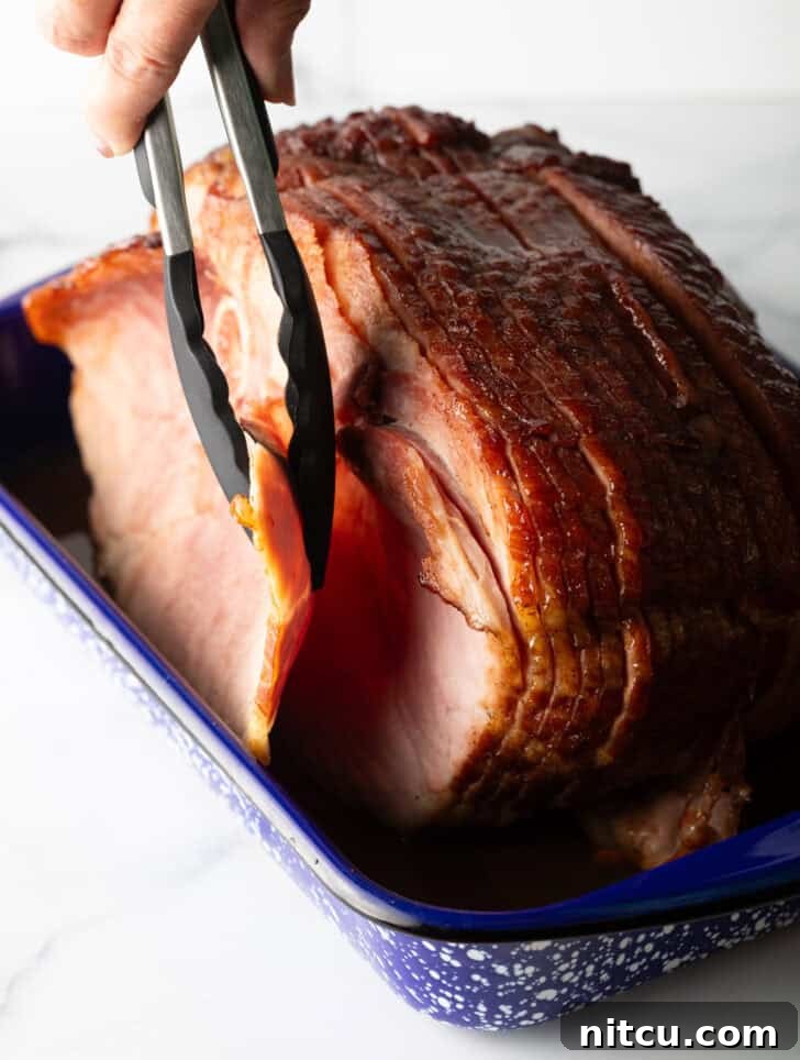 Perfect Spiral Ham Recipe 11 Tongs gently removing the front slice of a perfectly cooked spiral ham from a baking dish, showcasing its tenderness.