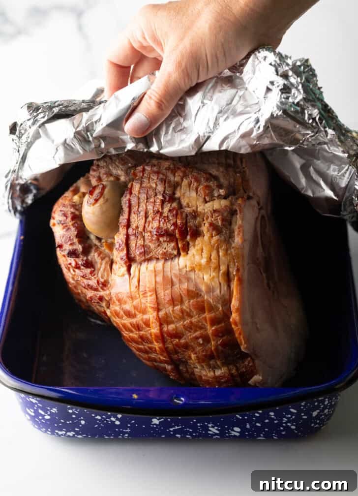 Perfect Spiral Ham Recipe 7 A cook carefully covering a spiral ham in a blue and white speckled roasting pan with aluminum foil, illustrating the step to lock in moisture.