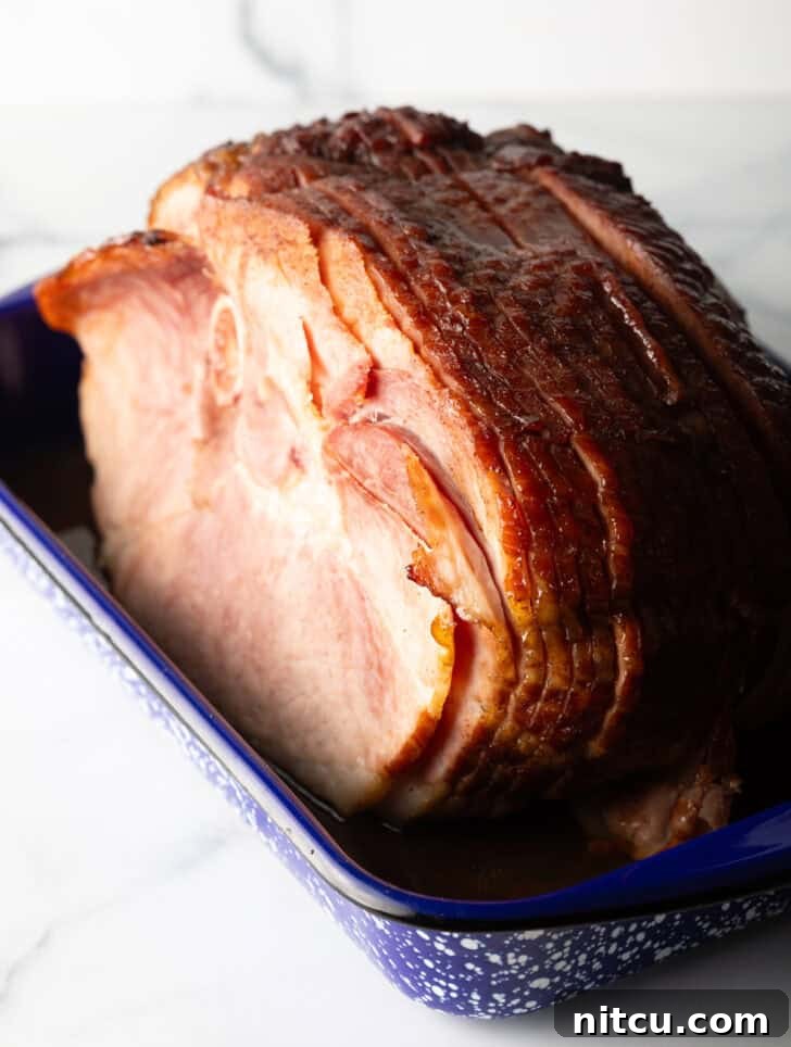 Perfect Spiral Ham Recipe 10 A cooked spiral ham, glistening with glaze, in a blue and white speckled roasting pan, with a few slices already carved.
