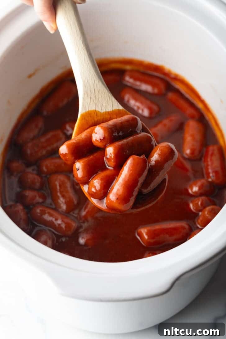 Glazed Cocktail Smokies 2 Wooden spoonful of smoked mini hotdogs in bbq sauce, held over the crock pot to camera.