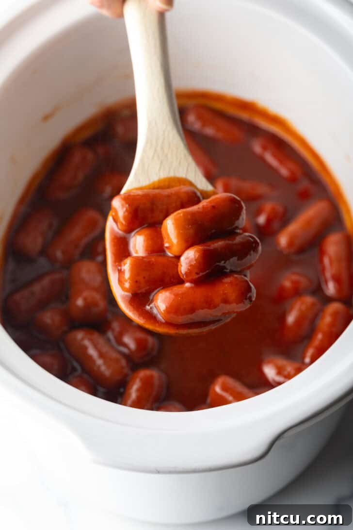 Glazed Cocktail Smokies 11 Wooden spoonful of smoked mini hotdogs in bbq sauce, held over the crock pot to camera.