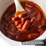 Wooden spoonful of smoked mini hotdogs in bbq sauce, held over the crock pot to camera.