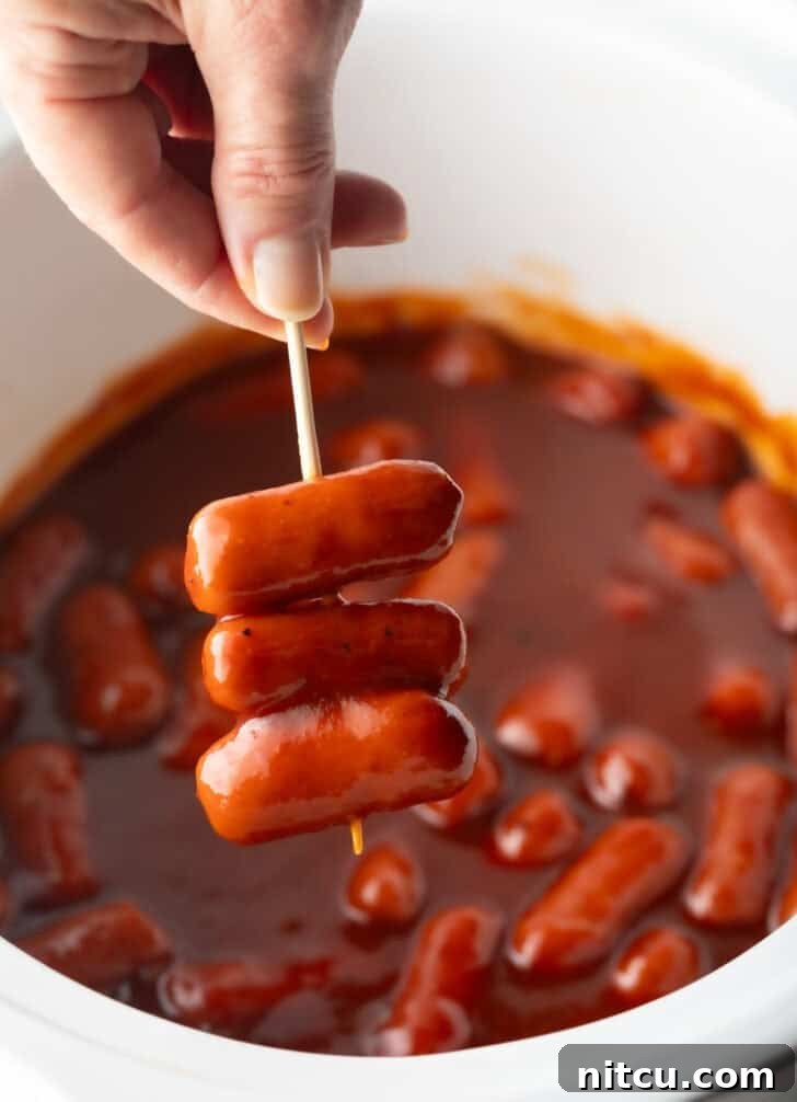 Glazed Cocktail Smokies 13 Hand holding a skewer with 3 little smokies, over a crock pot with cooked mini sausages in bbq sauce.