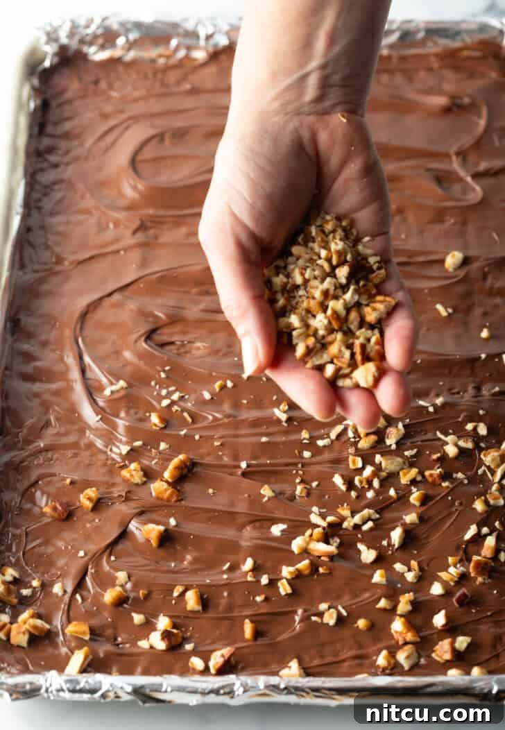 Hand sprinkling broken pieces of toffee overtop of chocolate covered saltine crackers in a baking sheet.
