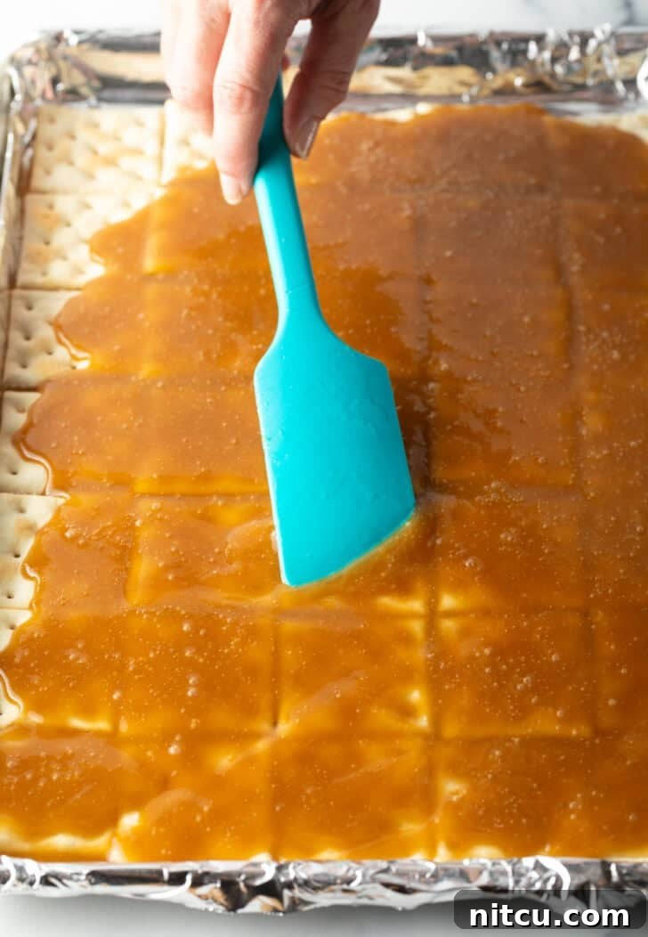 Using a blue spatula to smooth melted toffee across saltine crackers on a baking plan.