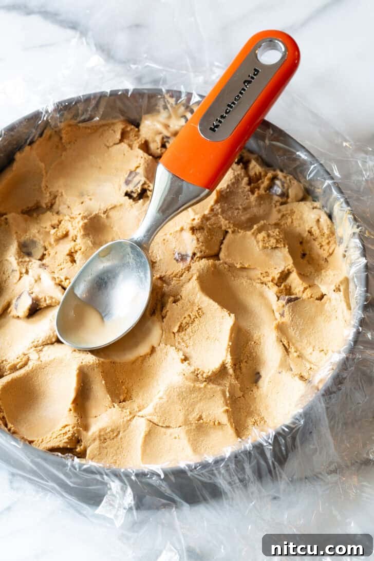 Ultimate Ice Cream Cake 6 Using a metal scoop to smooth ice cream into a cake pan lined with plastic wrap, to create a thick layer of ice cream.