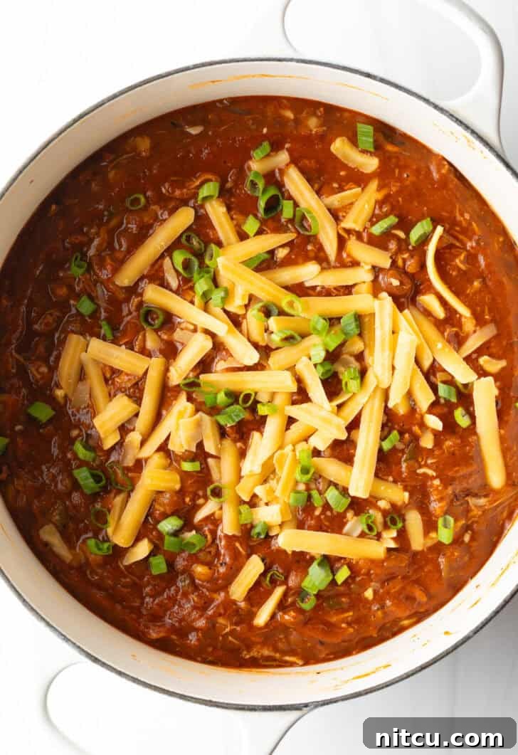 Easy 5 Ingredient Chicken Chili 12 A pristine white soup pot, seen from directly above, brimming with the inviting chicken chili, exquisitely garnished with finely chopped green onions and a generous layer of shredded cheddar cheese, presenting a visually appealing and comforting meal.