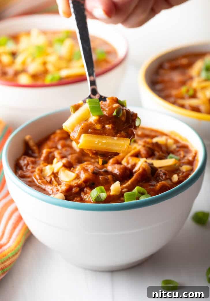 Easy 5 Ingredient Chicken Chili 13 A close-up view of a spoon digging into a white bowl filled with creamy shredded chicken, bean, and salsa chili, revealing the hearty texture and rich blend of ingredients within.
