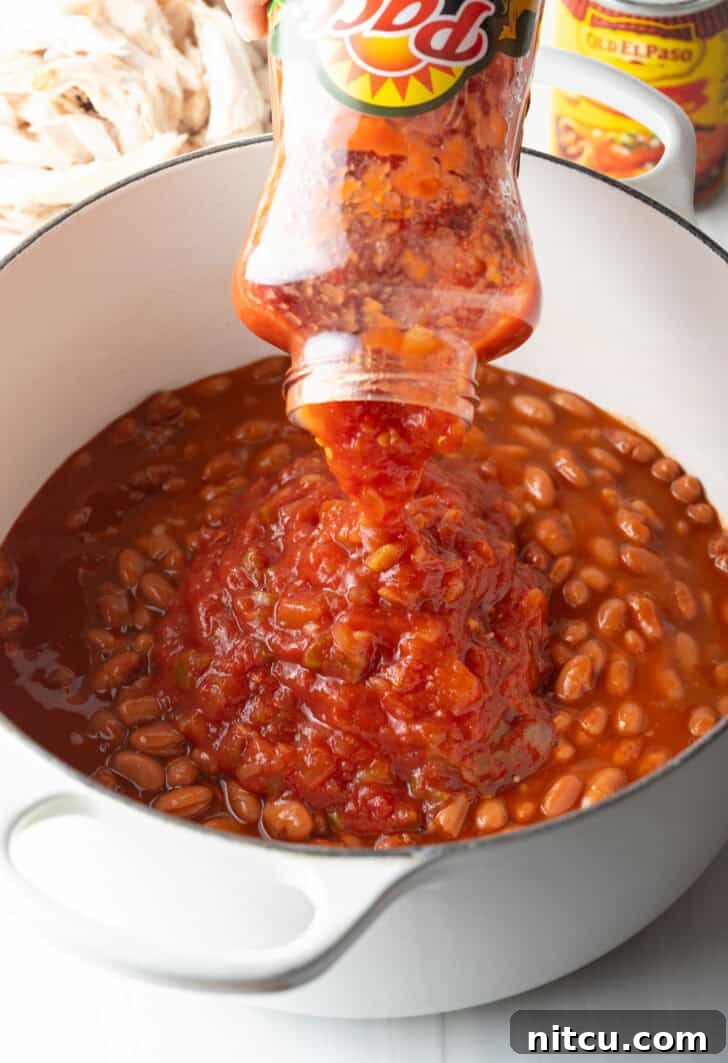 Easy 5 Ingredient Chicken Chili 7 A vibrant red enchilada sauce being poured into the pot, mingling with the pinto beans and creating a rich, aromatic foundation for the chili, capturing the essence of the 5-ingredient simplicity.