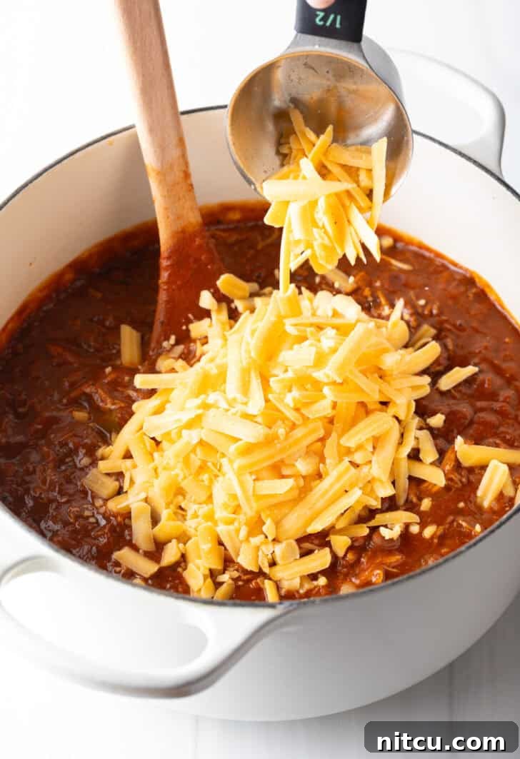 Easy 5 Ingredient Chicken Chili 9 A generous amount of shredded cheddar cheese being carefully added to the large pot of warm chili, poised to melt and create a creamy, luxurious texture that defines this easy chicken chili.