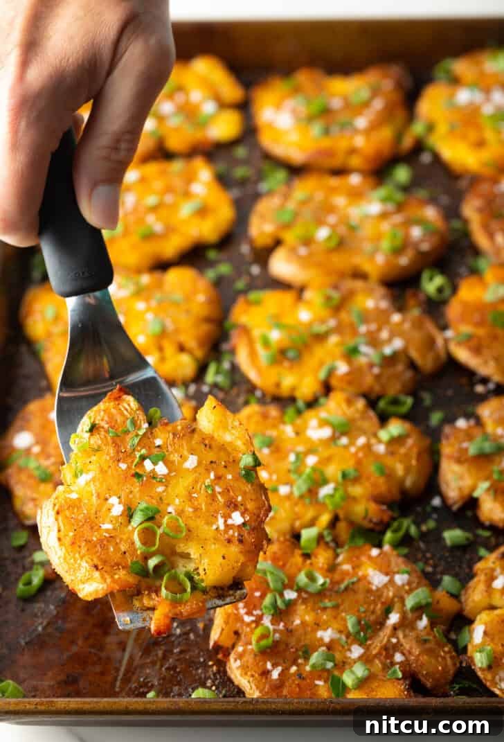 Crispy Smashed Potatoes 13 Top down view of 15 golden smashed potatoes on a baking sheet. A hand with a spatula is holding a crispy smashed potato to camera.