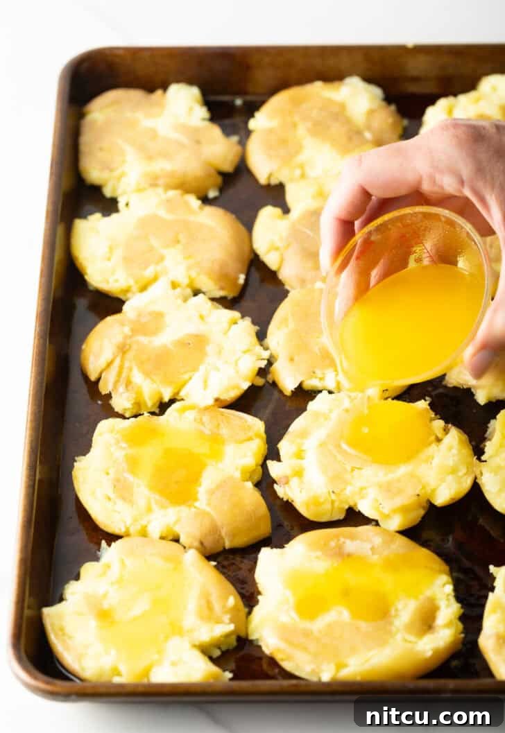 Crispy Smashed Potatoes 8 Hand pouring melted ghee from a small glass bowl over unbaked smashed potatoes on a baking sheet.