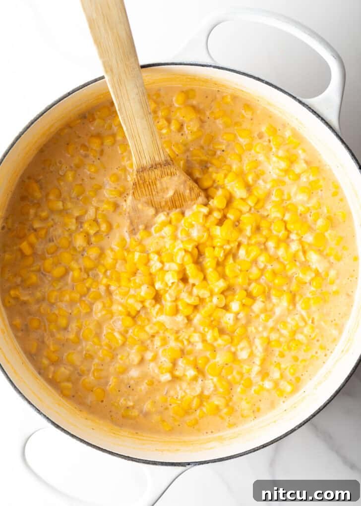 Top-down view of a wooden spatula stirring our creamy creamed corn recipe in a large white pot on the stove.
