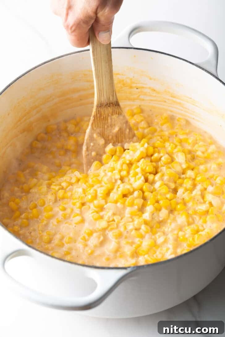 Wooden spatula stirring creamed corn in a large white pot, showing the rich, thick texture.