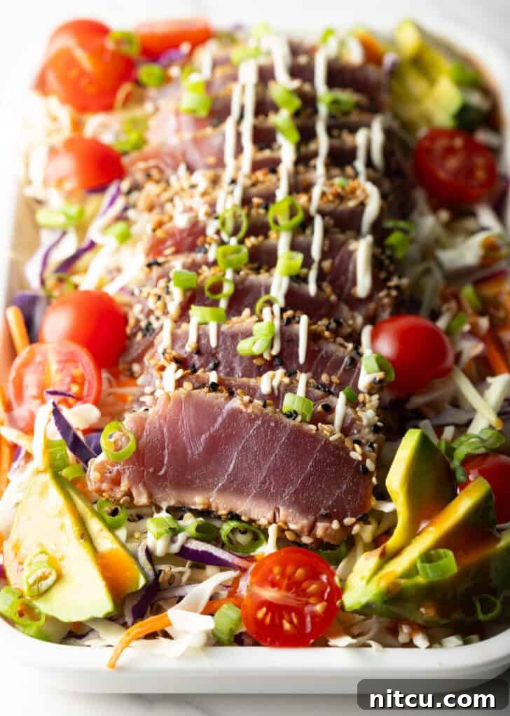 Flash-Seared Ahi Tuna 12 Large white rectangular dish loaded with fresh salad greens, halved cherry tomatoes, avocado slices, delicate slices of seared ahi tuna, chopped green onions, and a drizzle of dressing.