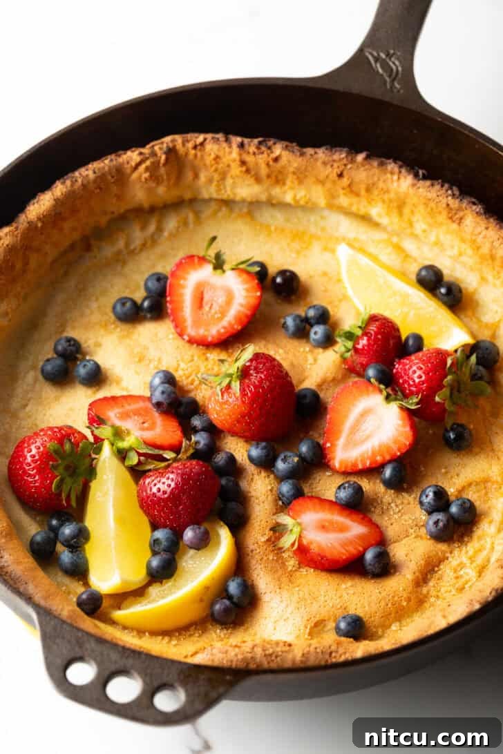 Effortless Puffed Pancake Perfection 8 A full Dutch Baby pancake in a cast iron skillet, garnished with an abundance of fresh strawberries, blueberries, and delicate lemon slices.