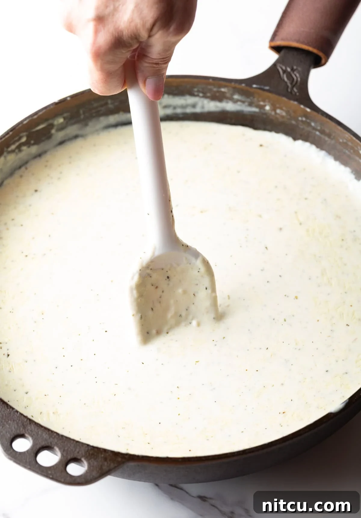 Velvety Chicken Alfredo Lasagna 6 A hand stirring creamy Alfredo sauce in a large stainless steel skillet with a white spatula.