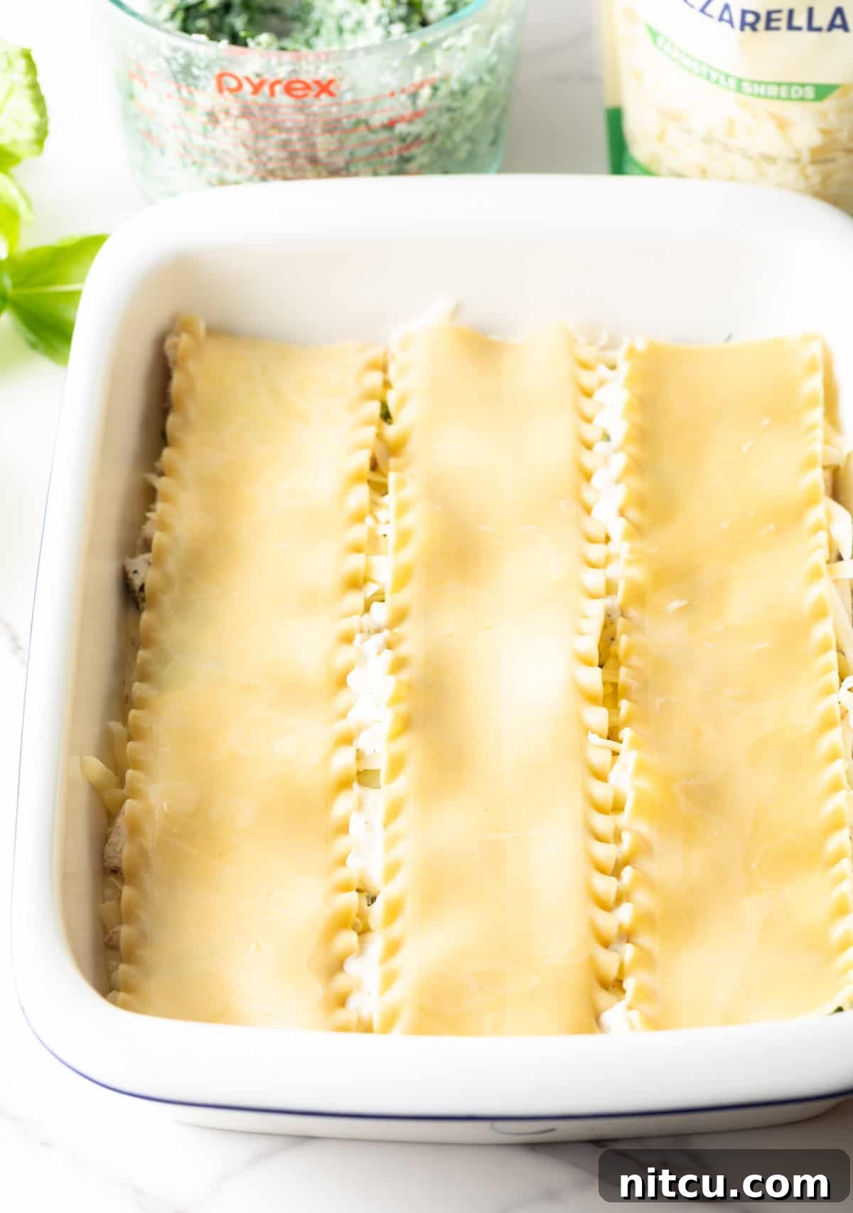 Velvety Chicken Alfredo Lasagna 8 A top-down view of lasagna noodles being carefully placed in a white rectangular casserole dish as the base layer.