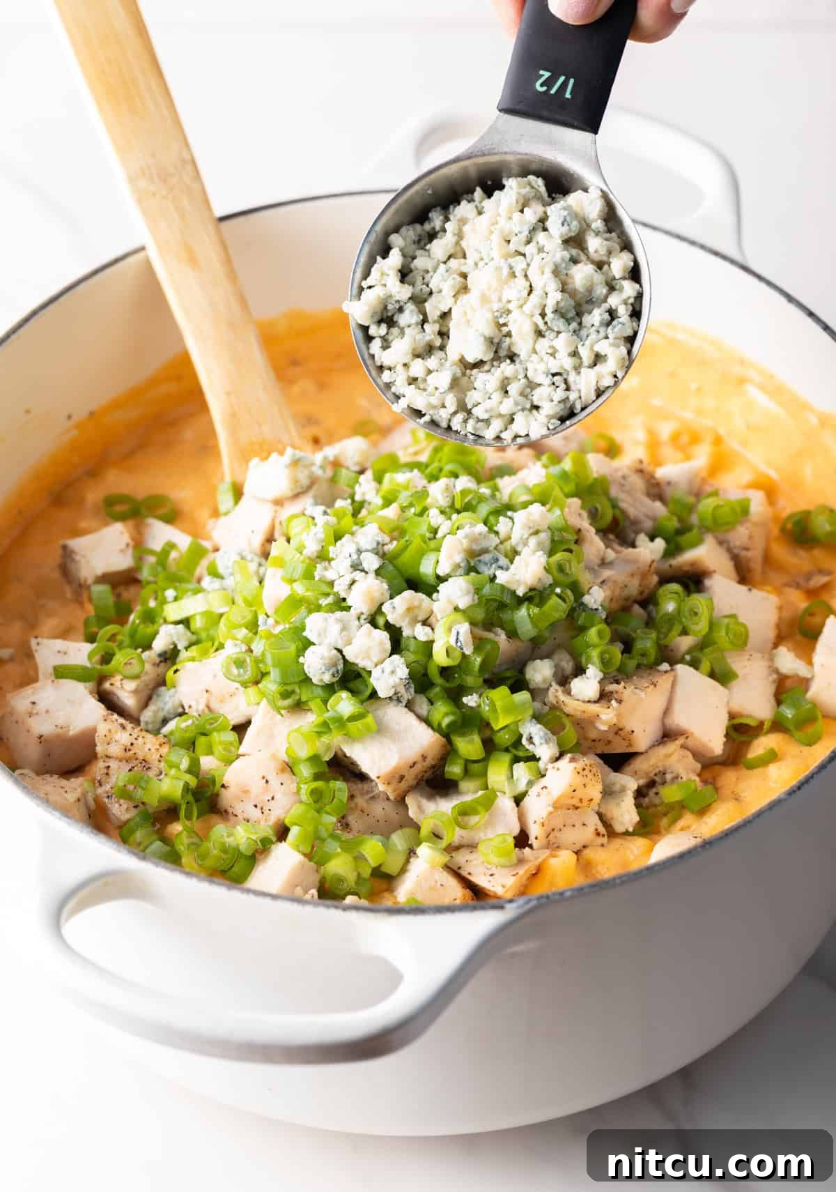Adding crumbled blue cheese from a metal measuring cup onto a mound of cooked chopped chicken and chopped green scallions in a large pot of macaroni and cheese.