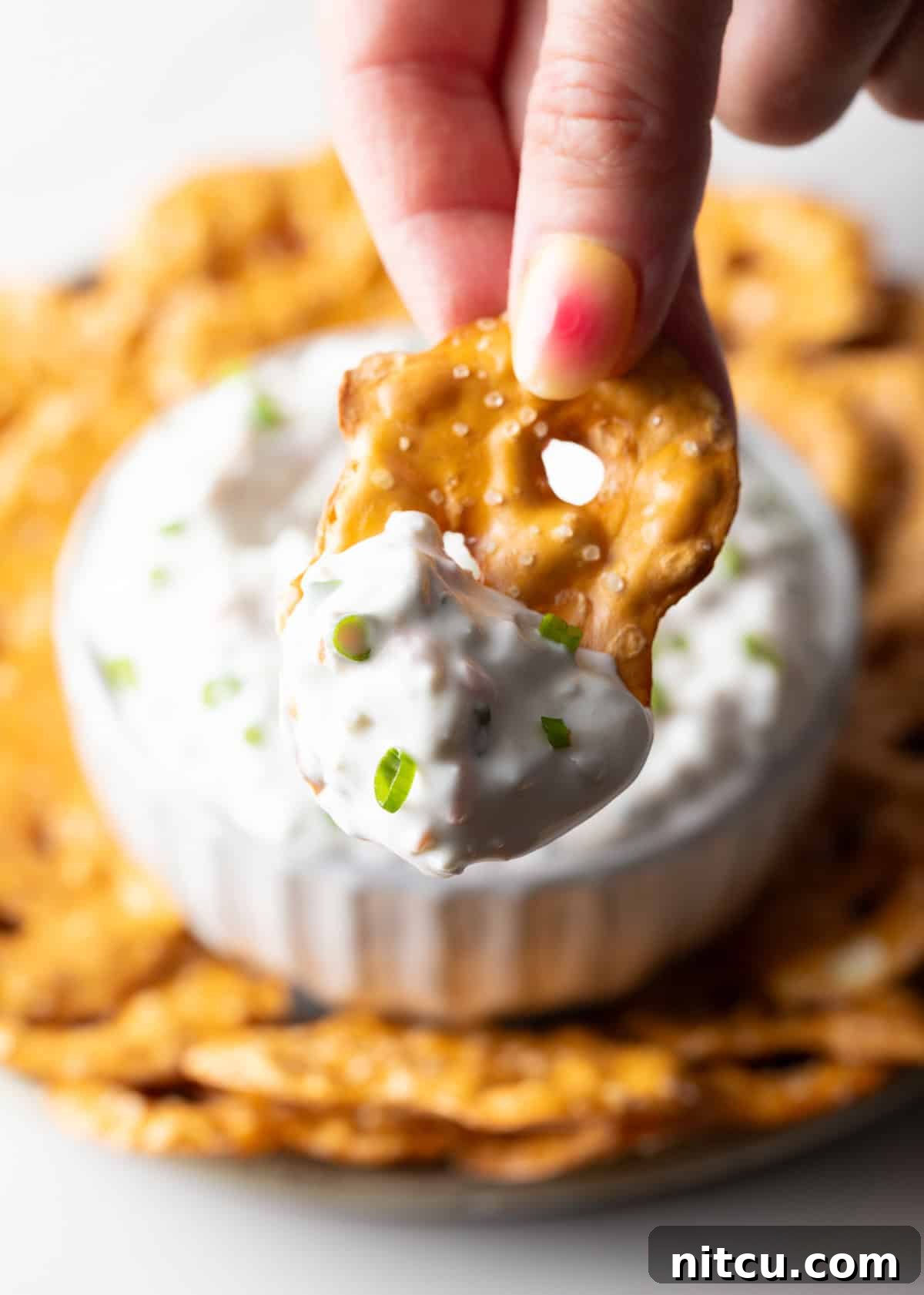 Hand holding a pretzel chip with a generous scoop of the creamy olive dip, ready to eat.