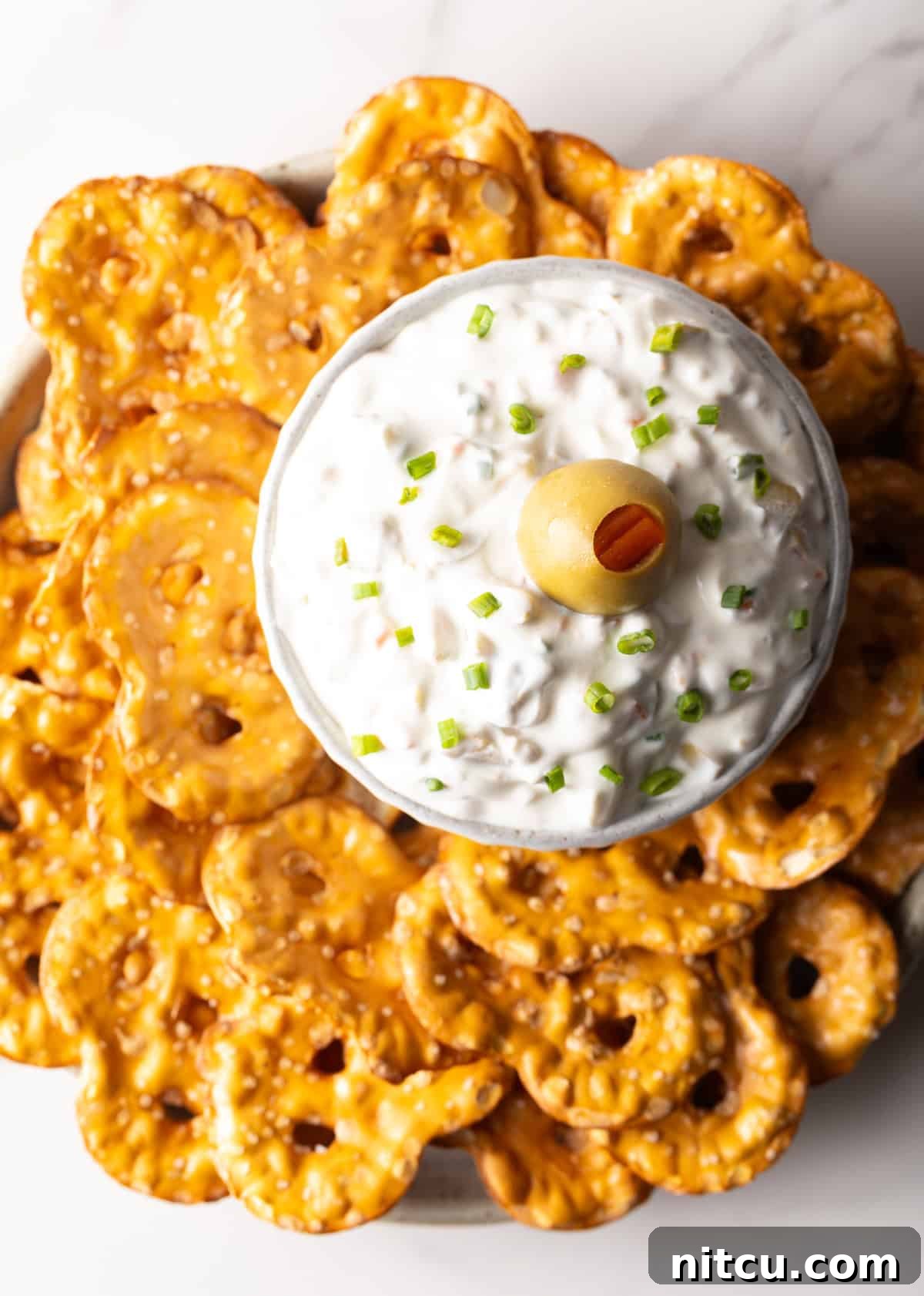 Top down view of a white bowl of dirty martini dip, garnished with a single green olive, and surrounded by crisp pretzel chips.